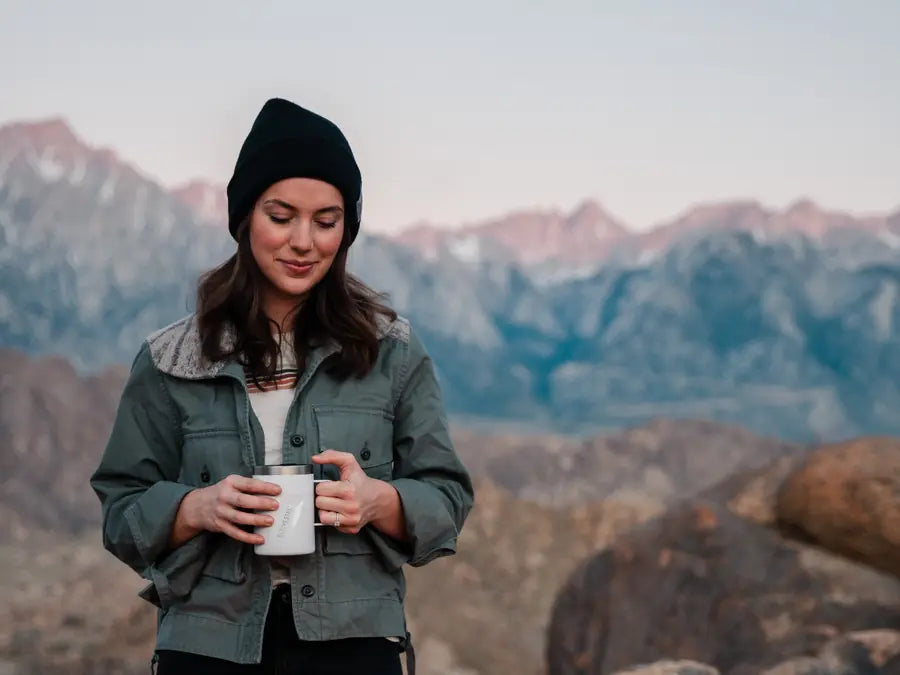 A woman in a beanie holding a white EcoVessel mug at dusk, mountains behind her